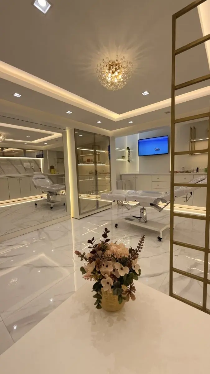 Visão ampla do consultório de biomedicina estética, exibindo a maca de procedimentos, mesa de planejamento facial e armários com portas espelhadas.